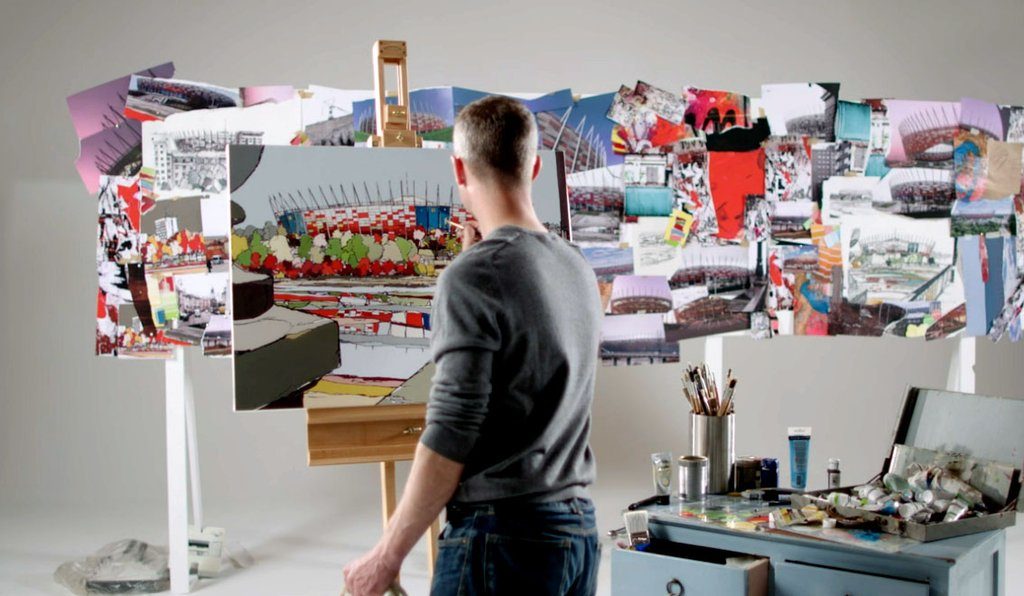 URBAN COLOURS ART REVIEW, PHOTO OF JAMIE THE ARTIST PAINTING A PICTURE WITH OTHER COMPLETED PICTURES AROUND HIM. HE IS STOOD UP WITH ART SUPPLIES TO THE RIGHT OF HIM