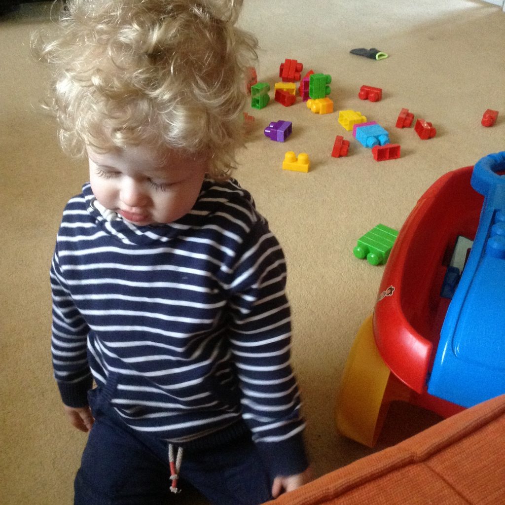Happy days Lucas in a blue and white striped hooded top, looking down with a slight smile and big blonde curly hair. Megablocks are scattered on the carpet behind him