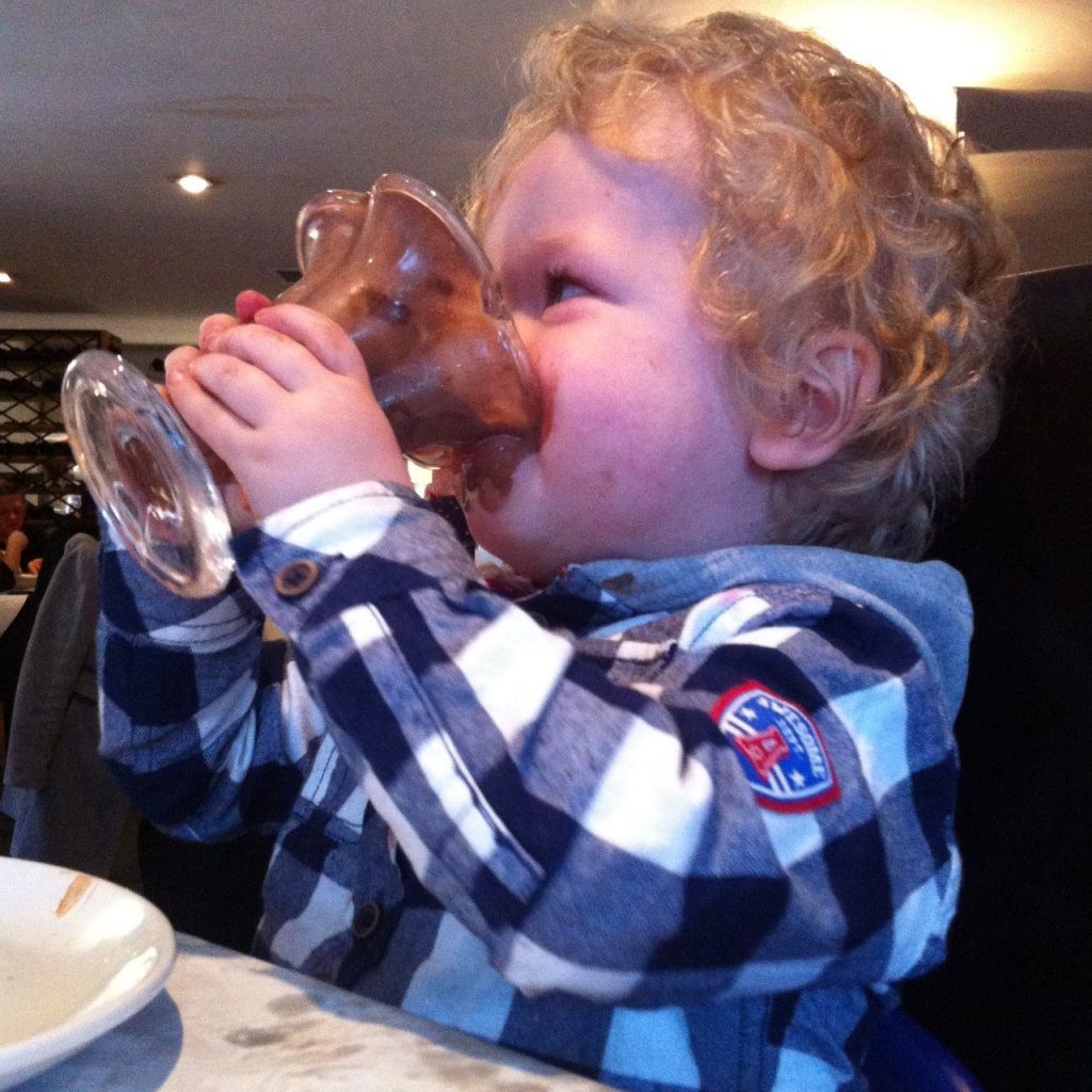 the ordinary moments Lucas drinking ,elted chocolate ice cream from a glass cup, he is wearing a blue and white checked shirt with grey hood, blonde curly hair