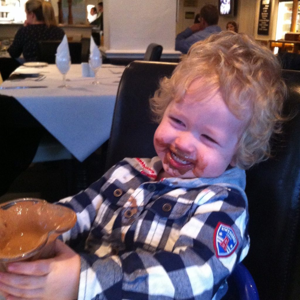 the ordinary moments Lucas sat smiling with cocolate ice cream all around his mouth. An empty glass of iceceam in front of him, his hands clutching it.he is wearing a blue and white checked shirt with grey hood, blonde curly hair