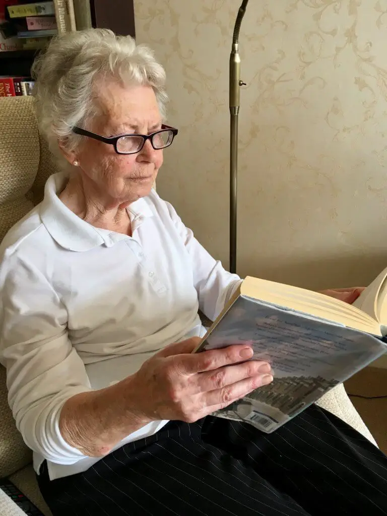 Eyejusters review My Grandma (an elderly lady with grey curly hair) is sat on a chair in a white top and black trousers on wearing purple glasses reading a thick hardback book
