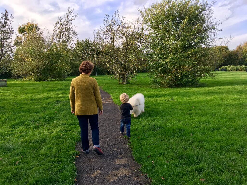 Walking the dog my mum and Lucas walking a big white fluffy dog through the field. My mum has a mustard coloured jumper in with dark blue jeans, short brown curly hair. Lucas has short wavy blonde hair holding the dogs lead and wearing dark blue jeans and black long sleeves top