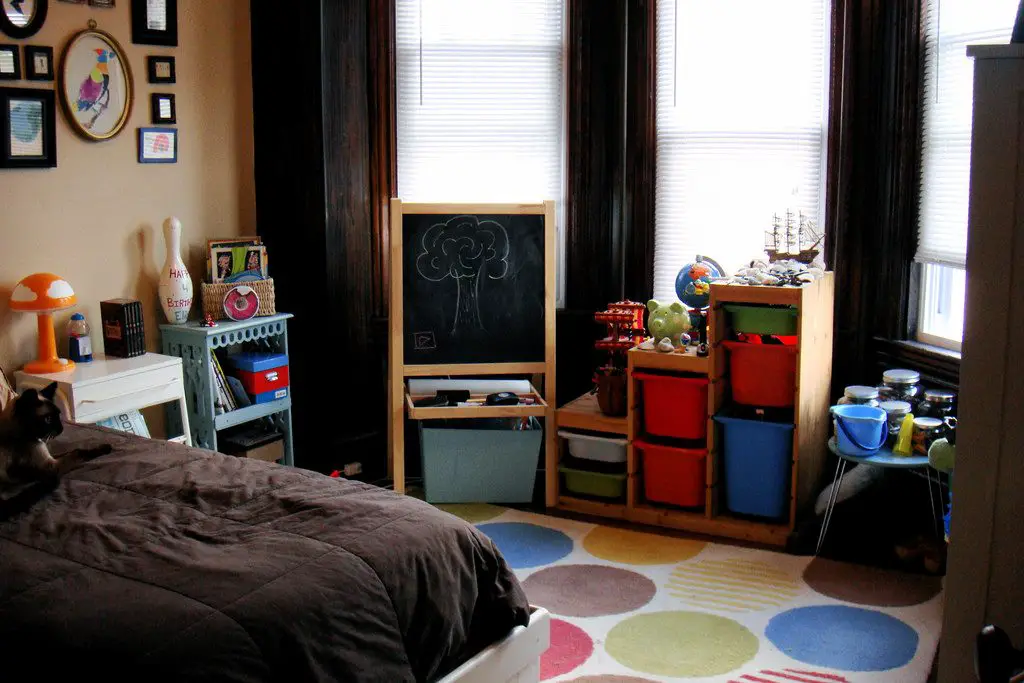 Choosing the perfect rug for your child’s playroom A bedroom with a bed showing brown bedding, a colourful spotted rug on the floor, a child’s chalkboard standing up and toys around the room
