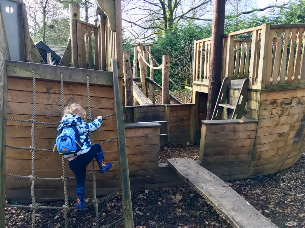 Samlesbury Hall, Preston Lucas is climbing up a rope ladder of the pirate ship wooden play area. He is wearing a blue geometric coat with a blue penguin backpack and navy trousers with blue wellies
