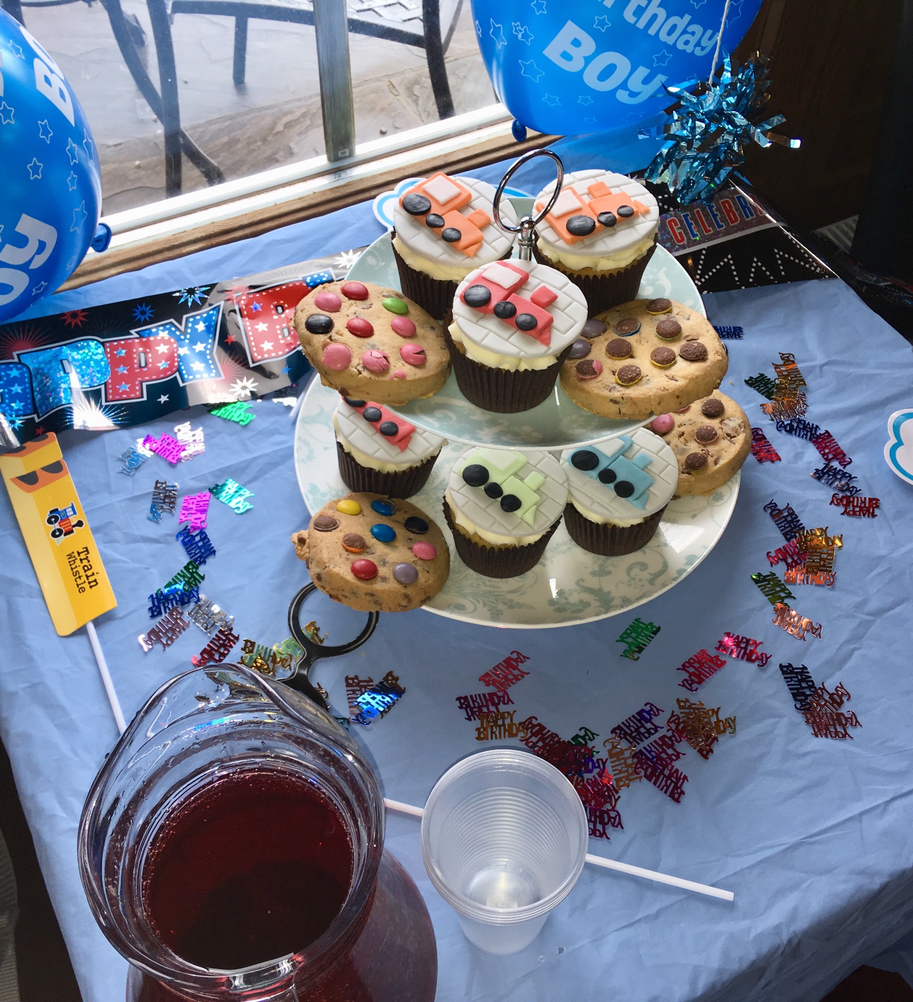 Lakeside Coffee House birthday afternoon tea a square table with blue paper covering and small colourful happy birthday decorations on. Blue birthday balloons and a 2 later stand of train decorated cupcakes and cookies decorated with smarties. A jug of black current cordial is in the corner of the table