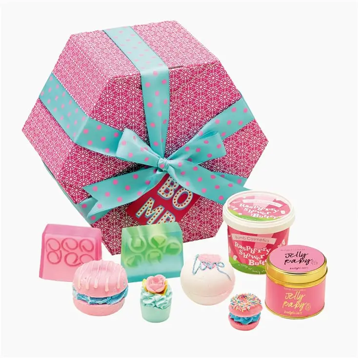 Mother's Day gift ideas a pink hexagon patterned box with blue ribbon and pink spots on, wrapped around the box in a bow. a selection of products in front of the box, soap, bath bombs and a candle