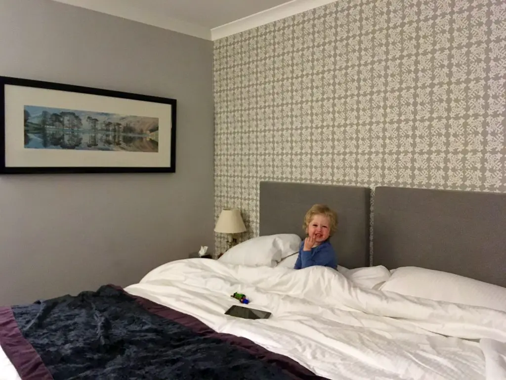 Low Wood Bay, Windermere review Lucas is sat in the super king bed covered with white bedding and wearing a blue pj top