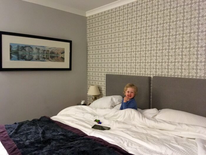 Low Wood Bay, Windermere review Lucas is sat in the super king bed covered with white bedding and wearing a blue pj top