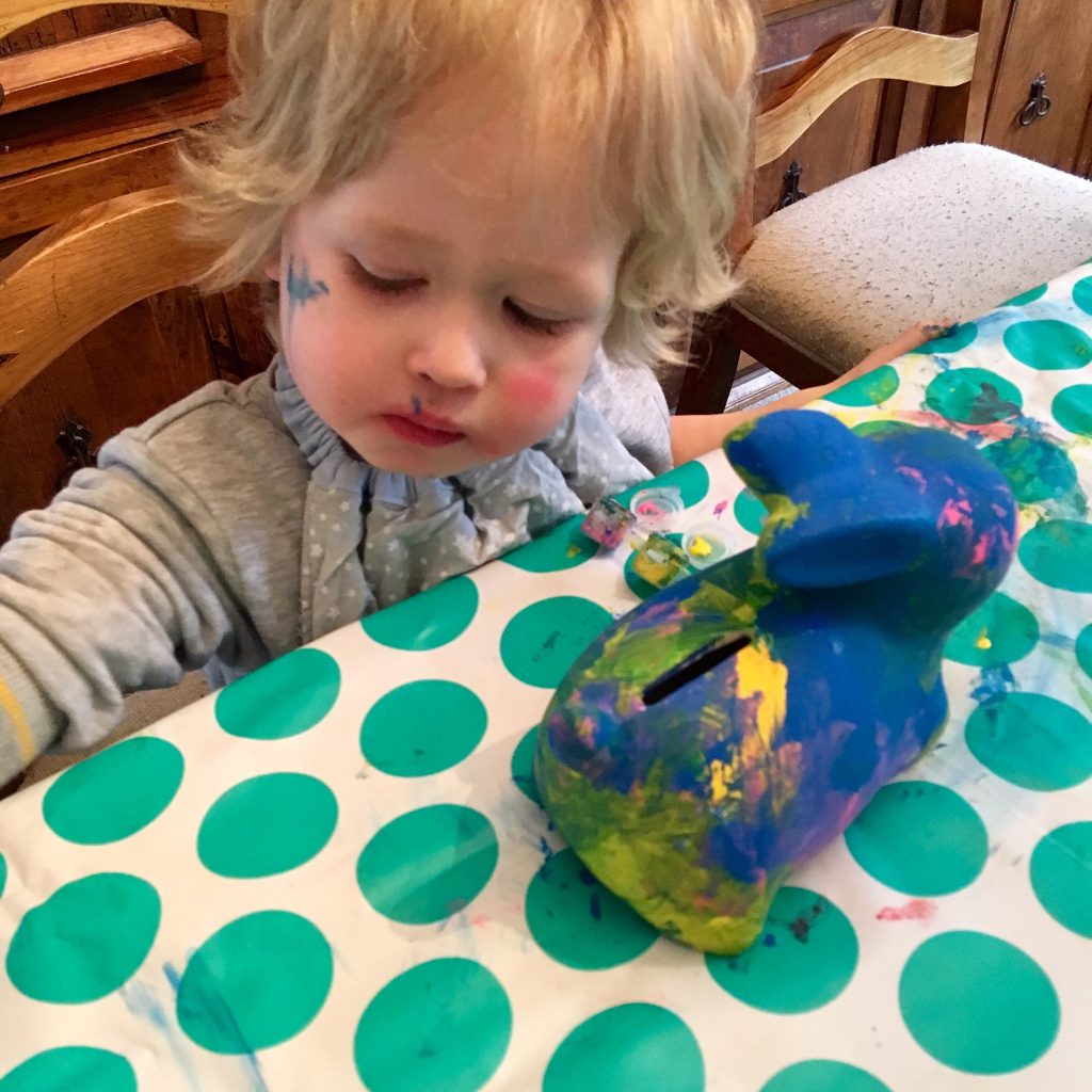 Easter 2018 Lucas is sat at the table with a white a green spotty mat, he is looking down and dipping his paint brush into paint whilst the bunny money box is partly painted next to him