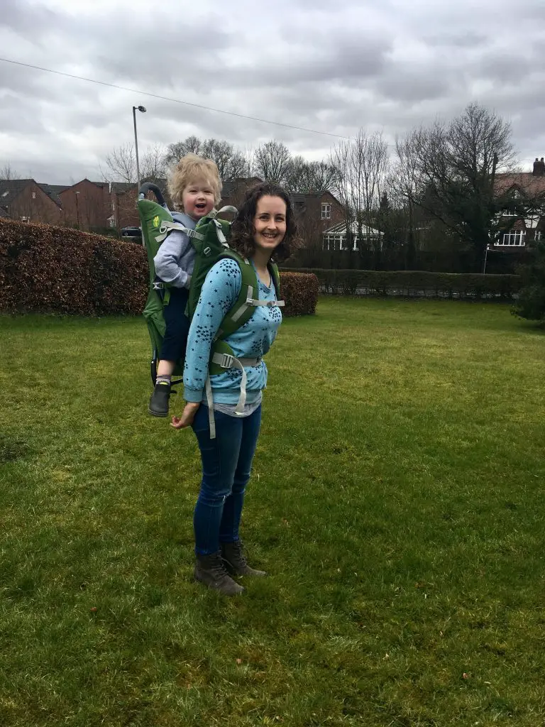 LittleLife Adventurer S2 review a photo of me standing in a field with my curly hair down, I am looking at the camera, wearing a blue and silver star print jumper and dark denim jeans, grey boots. I have the child carrier on my back and Lucas is smiling at the camera in the carrier. He has blonde wavy hair, is wearing a grey jumper and navy jeans 