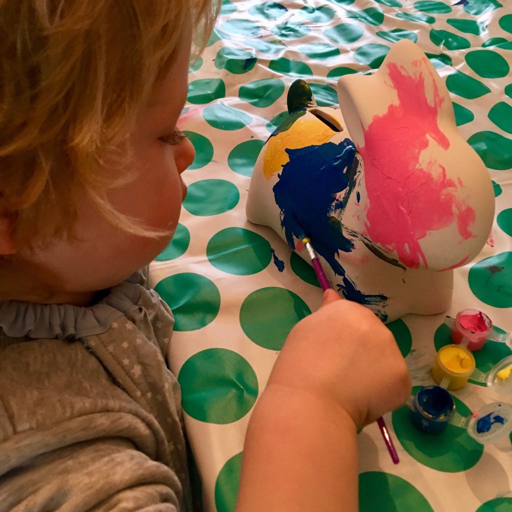 Easter 2018 Lucas is sat at the table with a green and white spotty mat under a white pot hubby rabbit money box he is painting blue pink and yellow