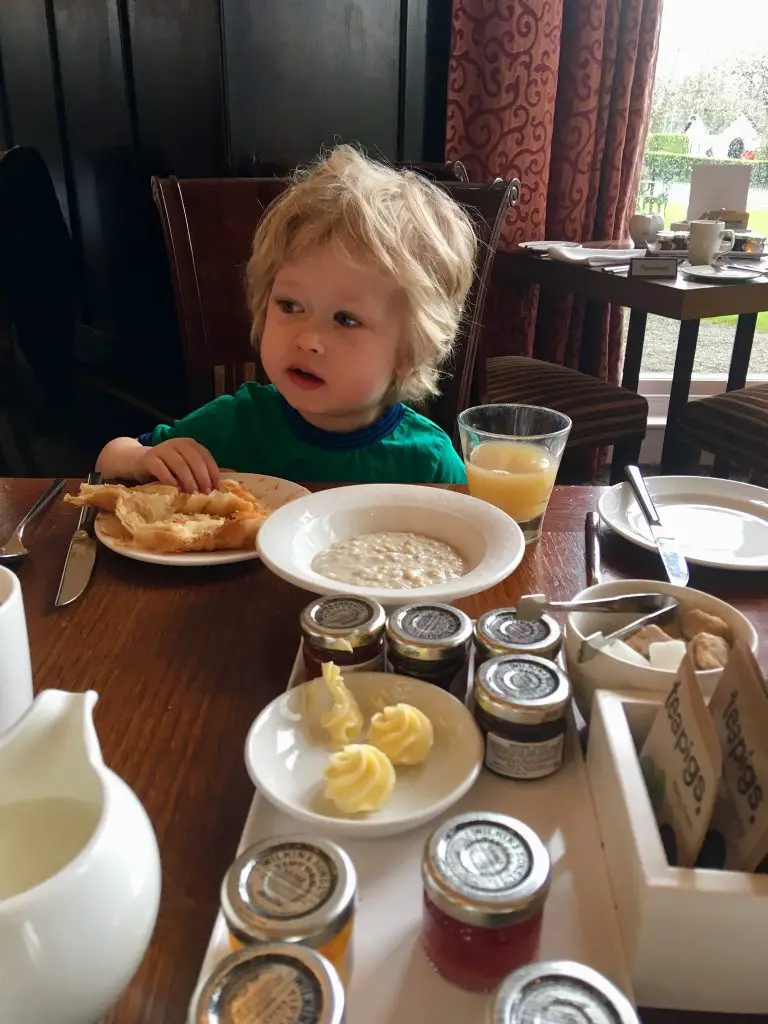 Low Wood Bay, Windermere review Lucas is sat a wooden table wearing a blue and green Tshirt. He has blonde messy hair. He is looking away from the camera. In front of him is a crossaint on his plate that his hand is touching. Before that is a milk jug and condiments