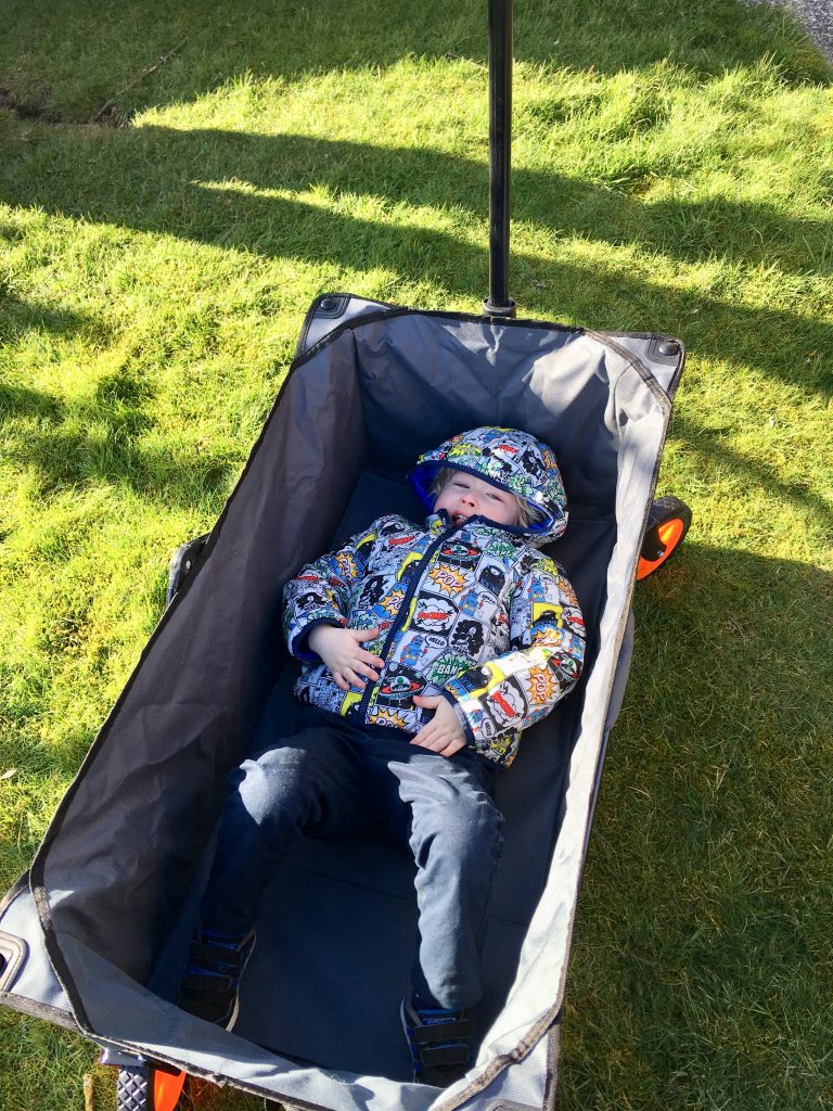 VonHaus camping folding cart review. The camera is looking down at Lucas lying in the camping cart. He has his hood up on his coat which is a superhero pow graphics over it. He is wearing navy pants and the cart is grey. Under the cart you can see grass. It is a sunny day