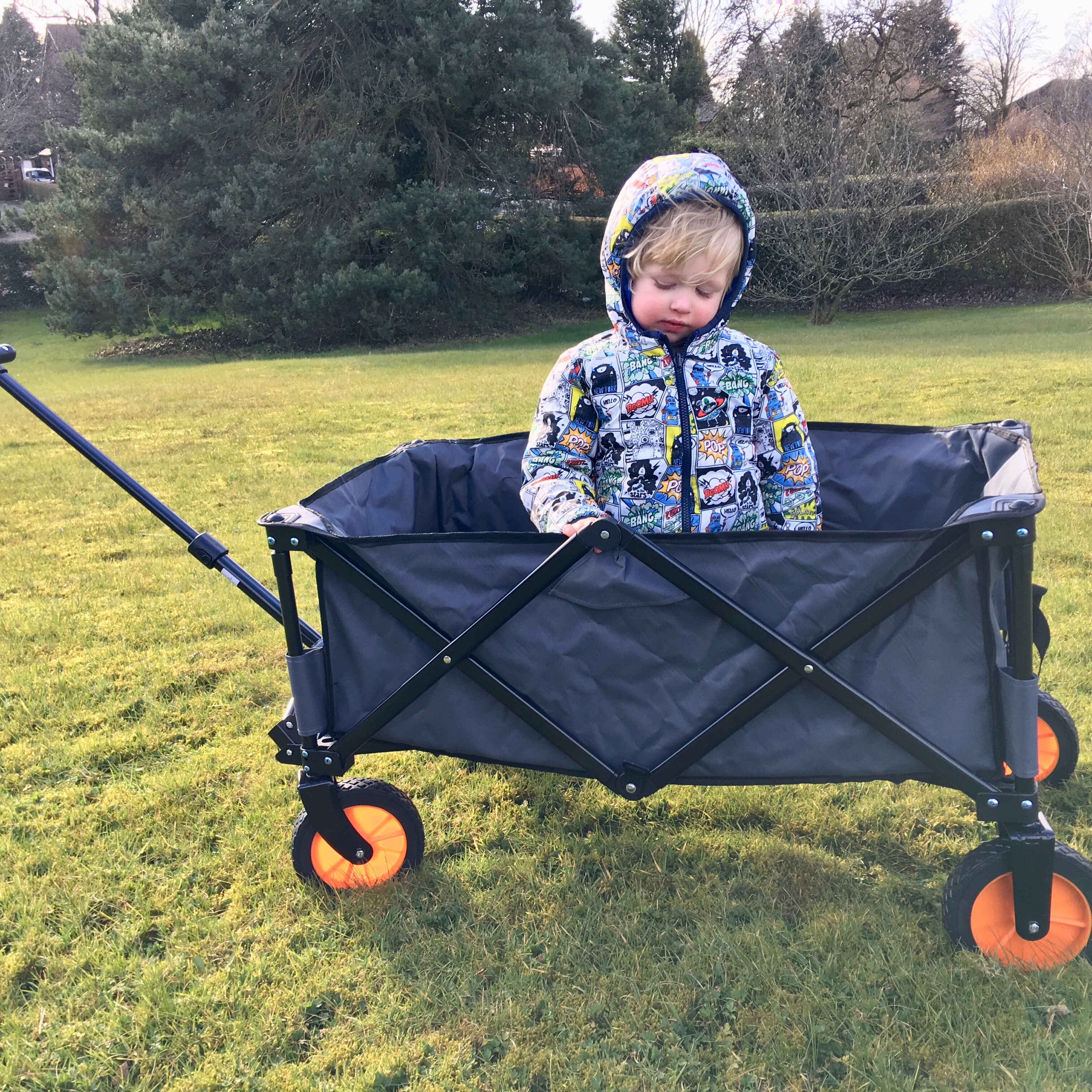VonHaus camping folding cart review. You can see the full side of the cart, it is grey with black steel frame and orange wheels/ Lucas is sat in the cart and you can see a field and tree in the background