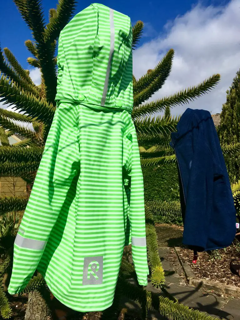 Reima jackets, 2 jackets hanging on a monkey puzzle tree in the garden with blue skies in the background. The jacket closest to the camera is a green and silver stripy rain jacket, the one further away in a navy fleece
