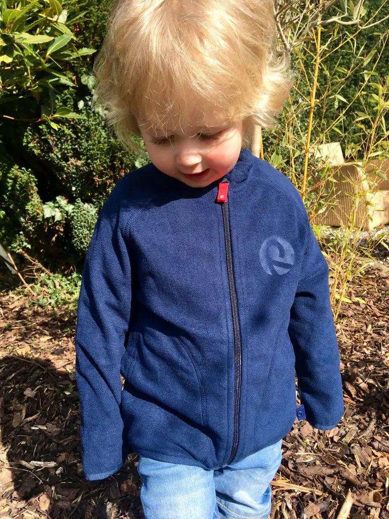 Reima clothing. Lucas is looking down smiling with blonde hair. He is stood in the garden wearing a blue fleece jacket