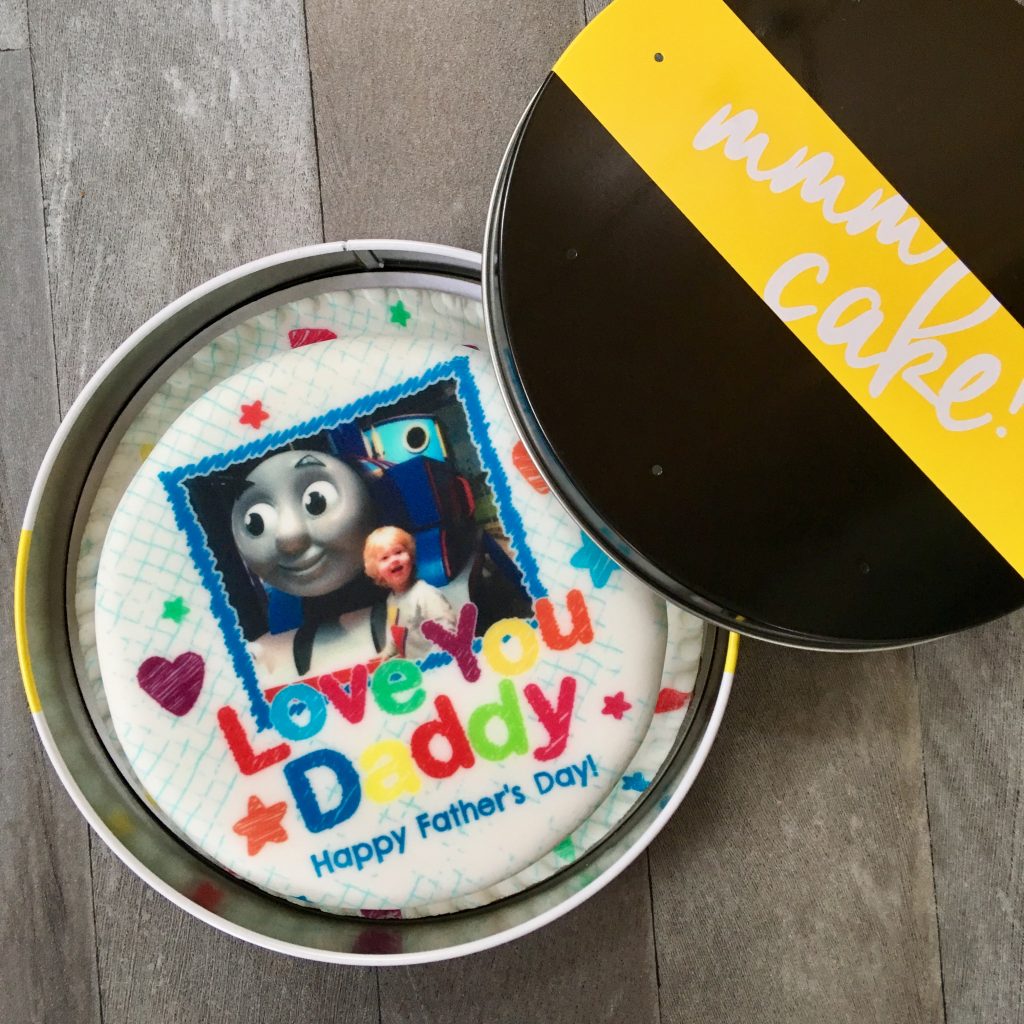 Father’s Day gift ideas a photo of a circular baker days cake in a tin. The cake has colourful writing saying happy father’s day and there is a photo of Lucas, a 3 year old blonde haired boy smiling at the camera next to thomas the tank engine