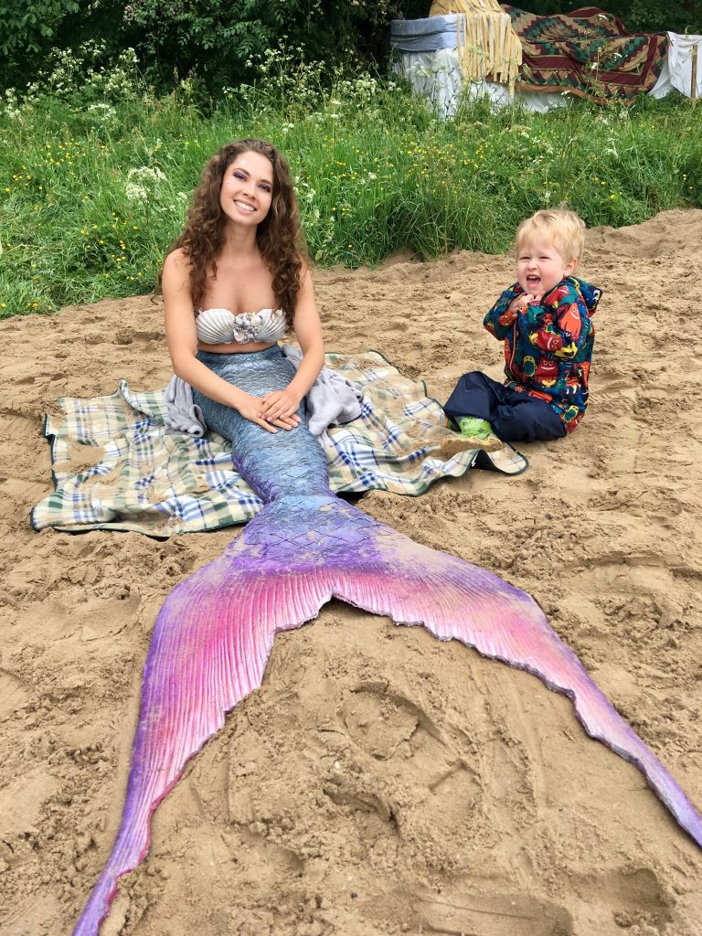 down b the riverside festival Lucas is sat next to a mermaid on sand, both smiling at camera