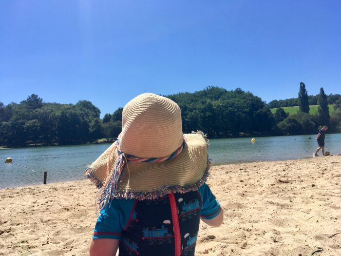 Summer essentials the back of Lucas who is wearing an all in one thomas tank engine sun suit and my straw hat looking out onto a lake day on sand