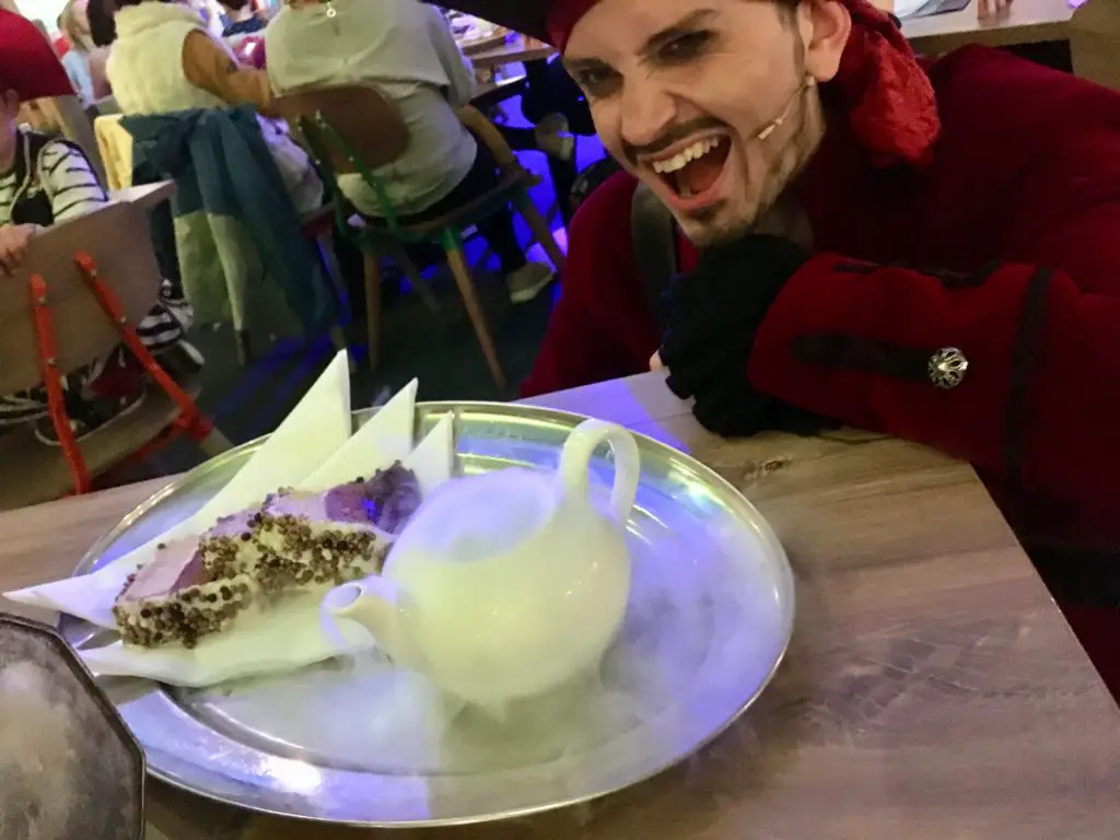 The pirate crouching down next to a silver tray of cake with a steaming tea pot 