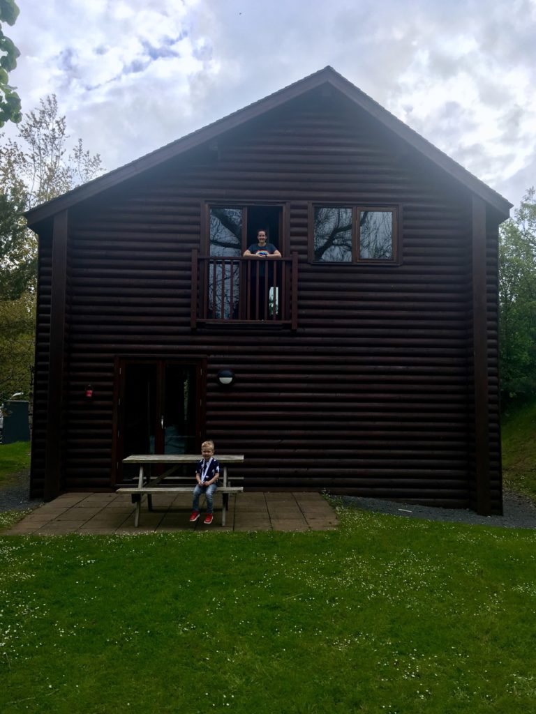 The dark brown wooden lodge. I am in the patio doors on the top level and Lucas is sat on the picnic bench outside 