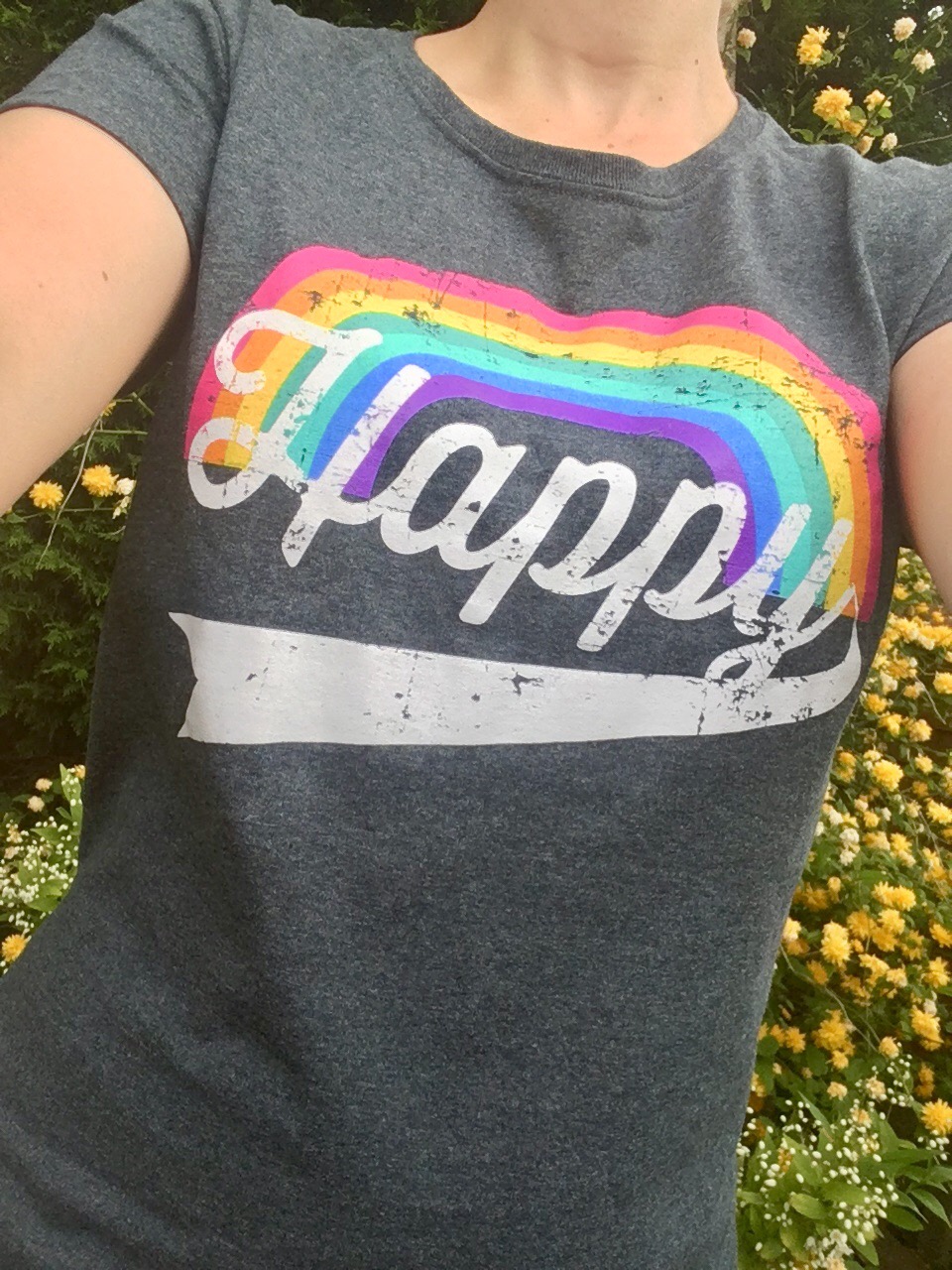 Close up of the charcoal T-shirt with a rainbow on and white Happy writing. Behind me are yellow flowers