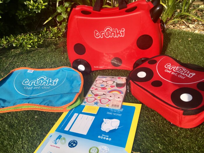 Travel with Trunki. The red and black spotted Trunki is on grass with the stickers, lunch bag and blue tidy bag next to it