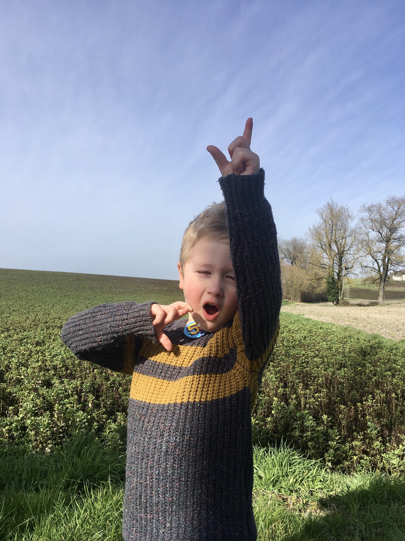 Lucas outside with one hand up, mouth open and eyes closed. Wearing a jumper and 5 badge