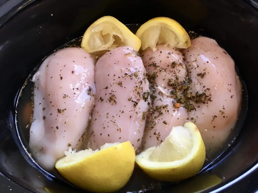 The chicken In the slow cooker with the stock poured over, chicken is covered in the herbs and the lemon rind is also on top