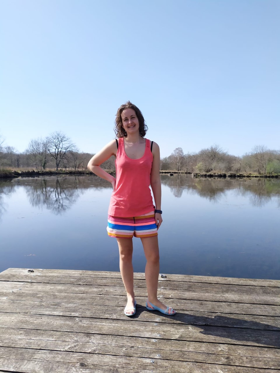 I am stood weraing a coral top and stripy shorts next to a lake