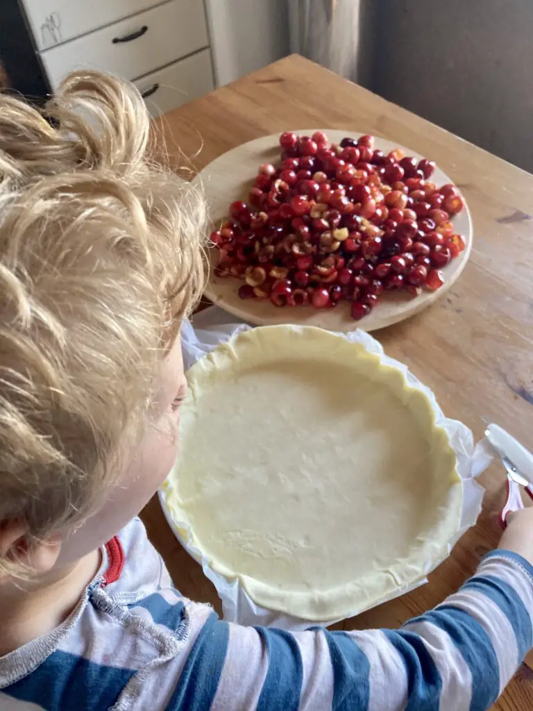 Lucas at the kitchen table with the pastry in a dish ready to put cherries in. The pitted cherries are on a board in front of him