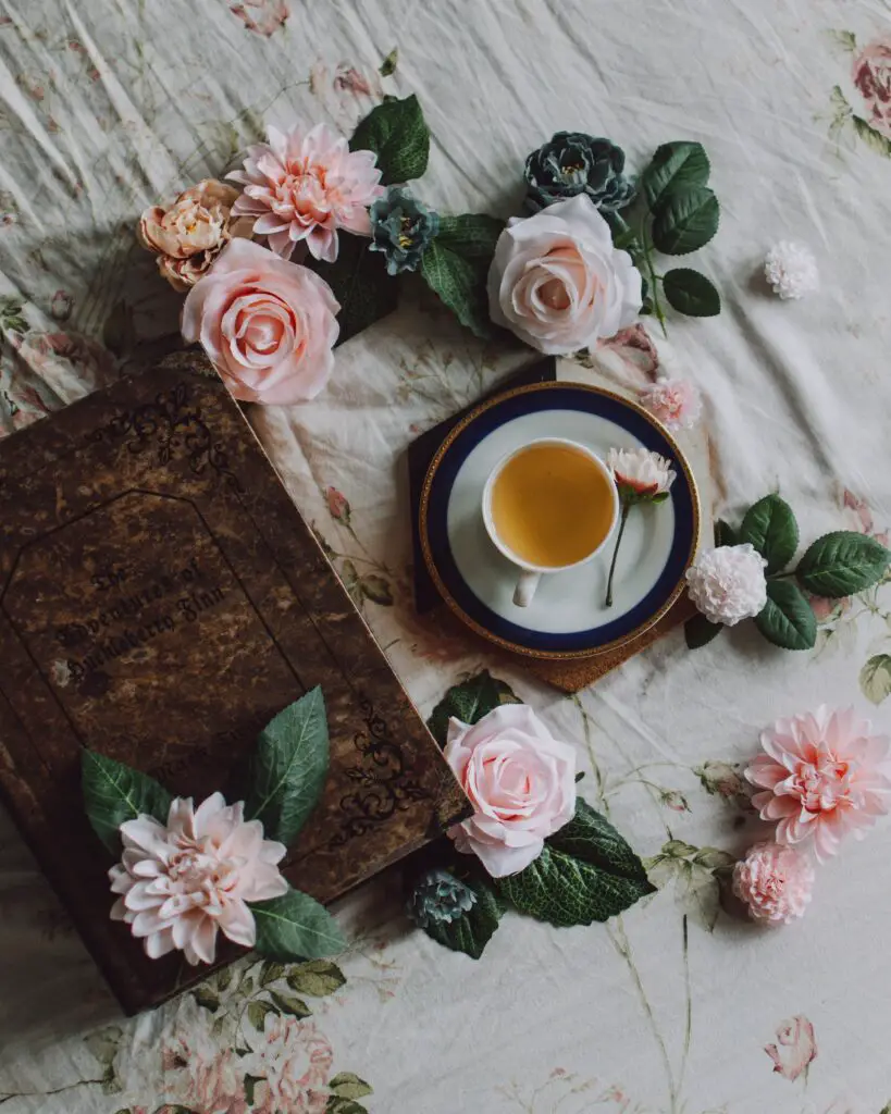 A photo taken from above. You can see a cup of white tea next to a brown book and pink flowers 