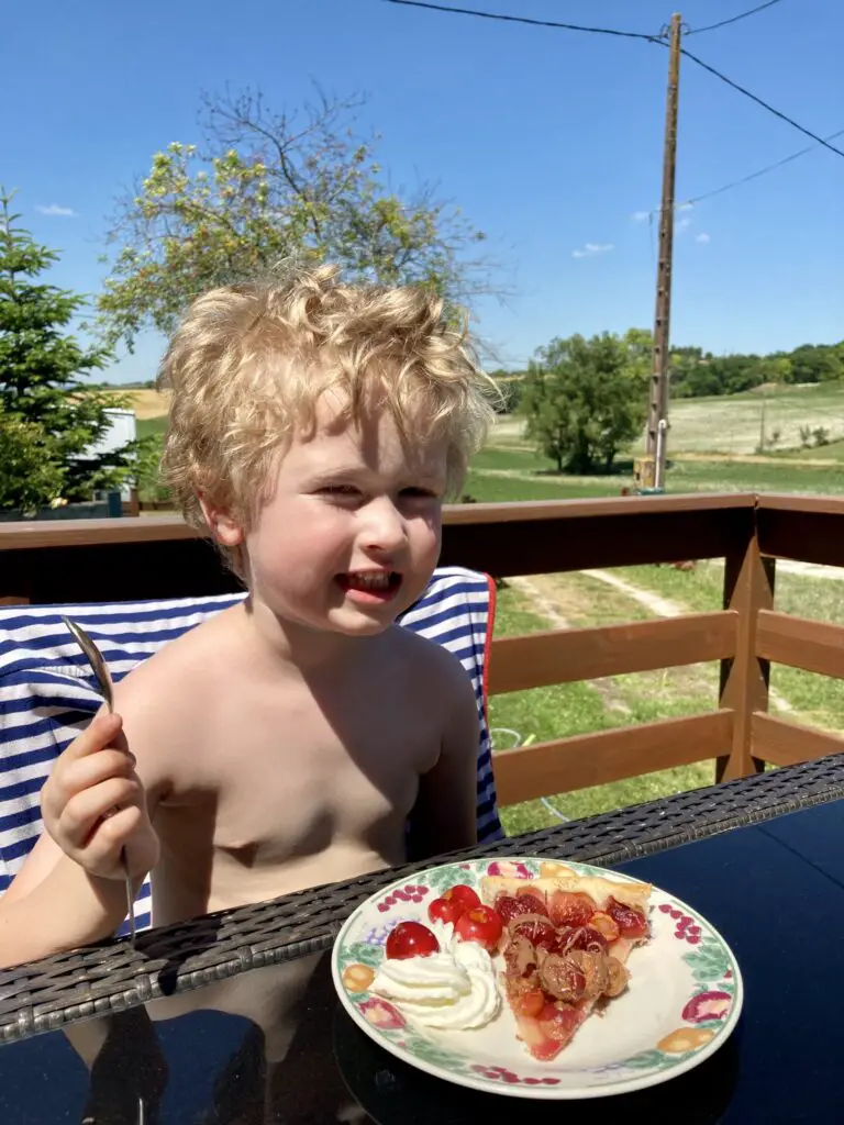 Lucas sat outside at the table smiling at the camera with a spoon in his hand and cherry tart in front of him 