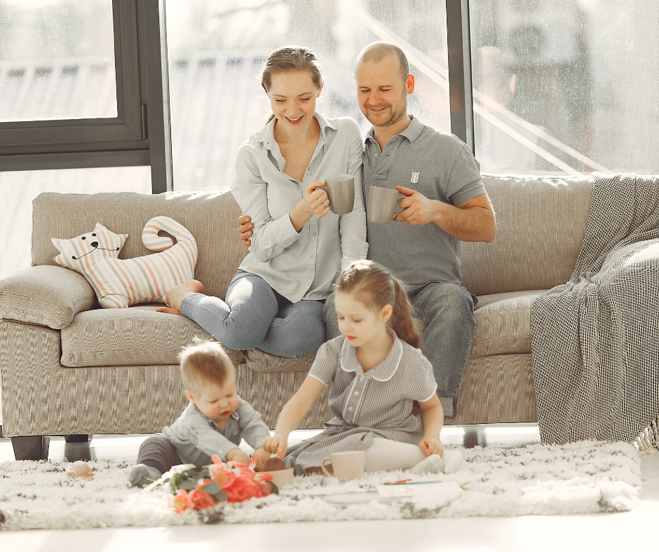 a family sat in the living room. the man and woman are sat on the sofa while a boy an dgirl play with toys on the floor