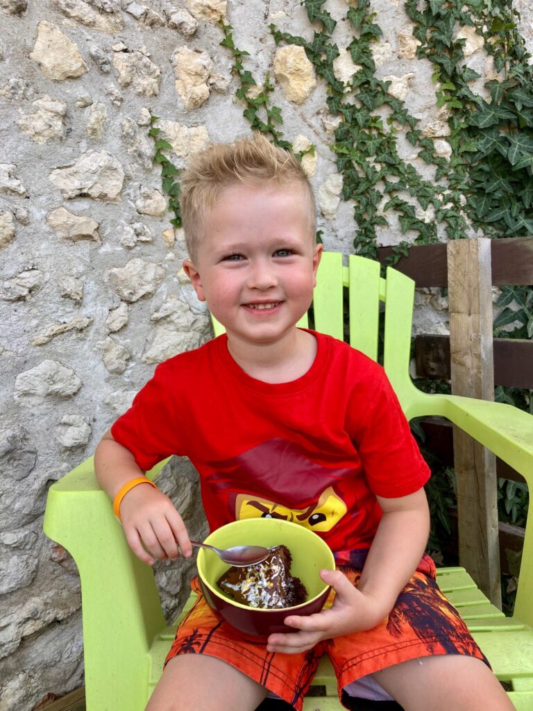 Lucas sat outside smiling at the camera. I’m his hands is a bowl with chocolate and hazelnut cake in with cream drizzled over 