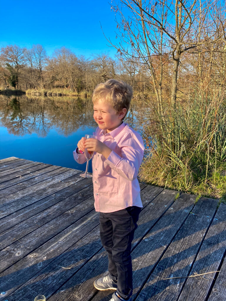Lucas is stood on decking with a lake behind him. He’s wearing a pink shirt and navy trousers. He holding a glass and smiling 