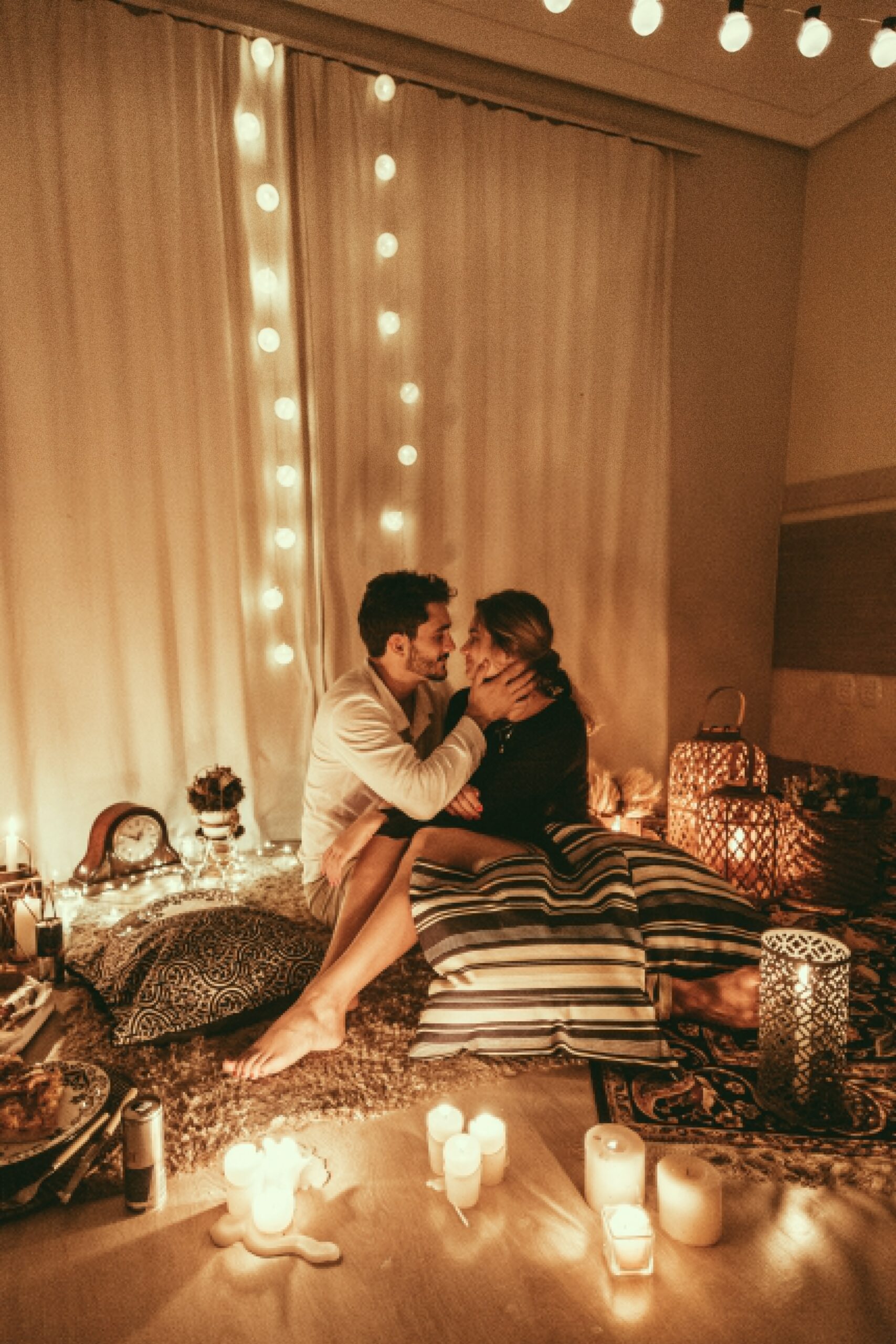 A romantic scene of a lady sat on a mans knee, they are surrounded by cushions and candles
