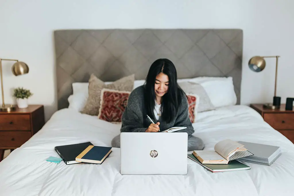 woman sitiing on her bed on the laptop with books around her