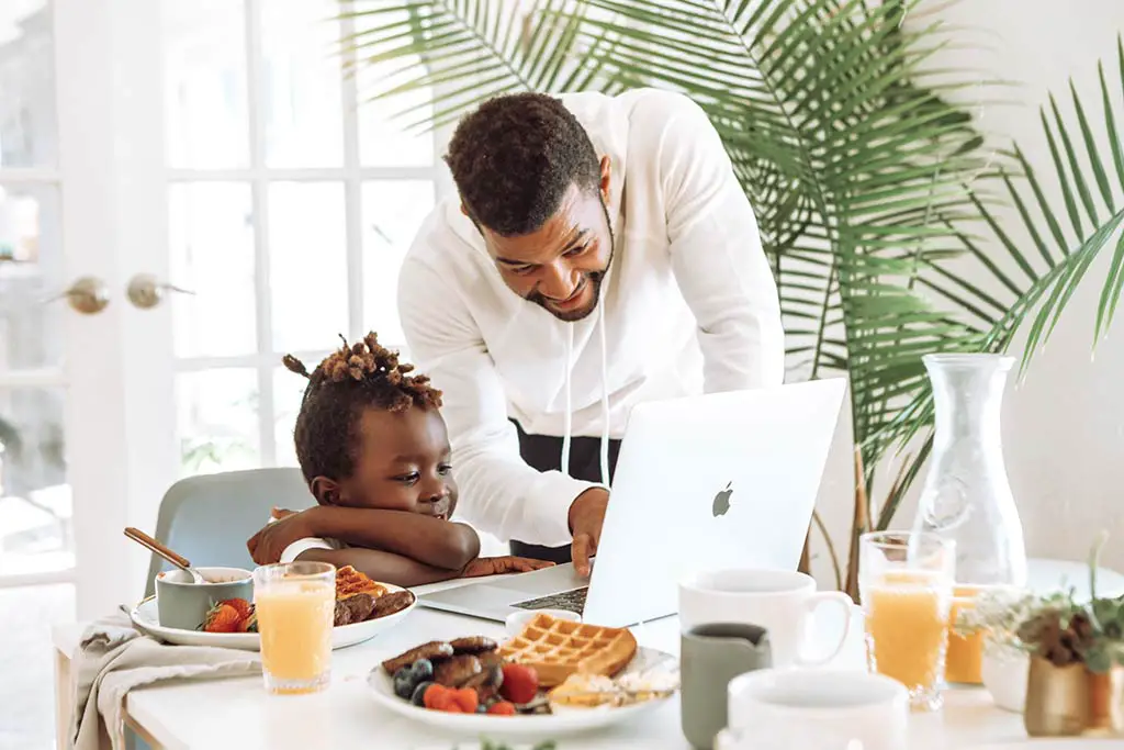 man at the dining table with chold pointing at the laptop with breakfast things on the table