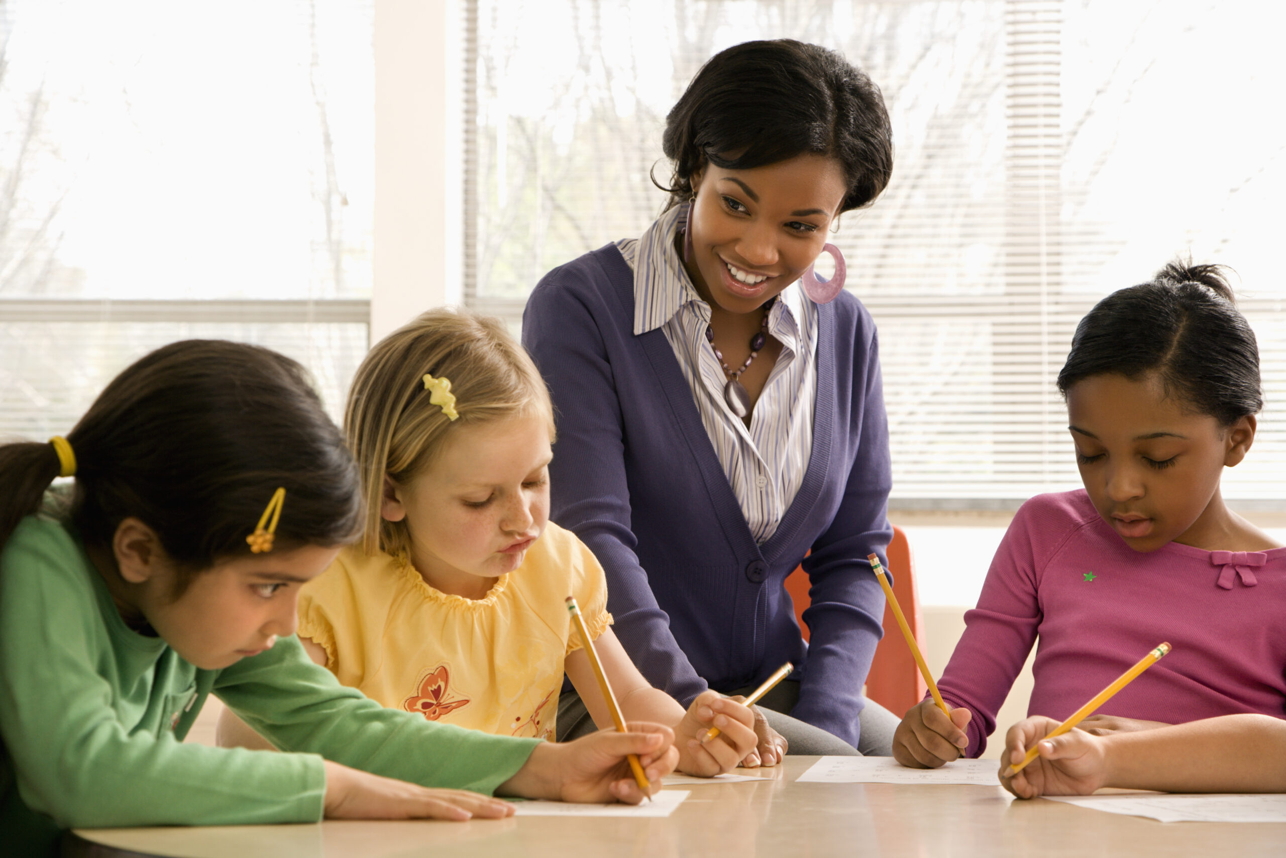 three children wiritng at a desk with an adult sat smiling at them. Teacher helping students in school classroom. Horizontally framed shot.