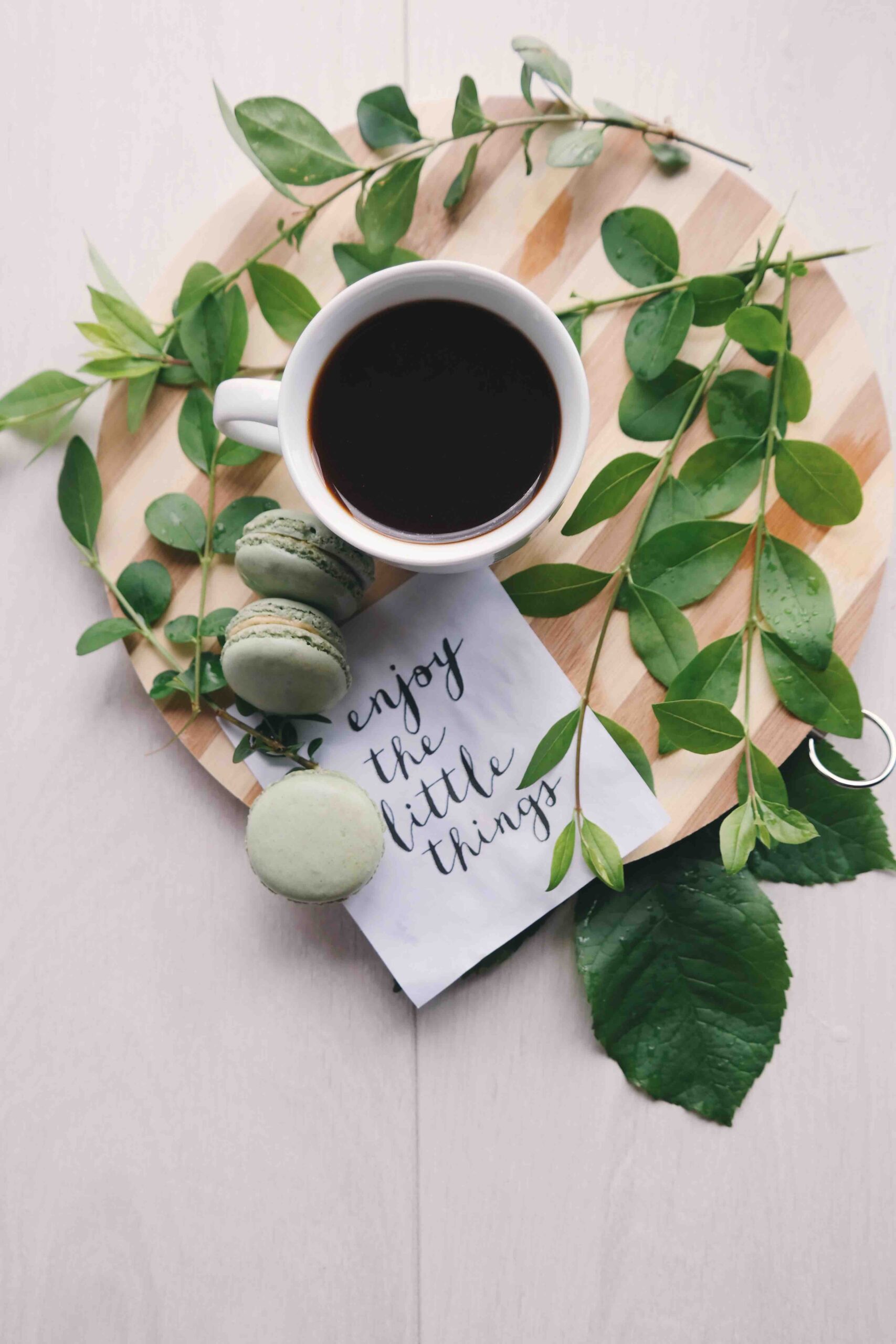 black coffee on a wooden board with leaves around and green acaroons next to a piece of paper with itallic writing enjoy the little things
