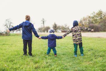 three children stood in a field holding hands with their arms outstreched, they are wearing outdoor winter clothes