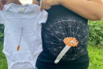 The photo shows a close up of my pregnancy bump with the dandelion design on the tshirt, I am also holding up a white baby grow that has a dandelion seed on it