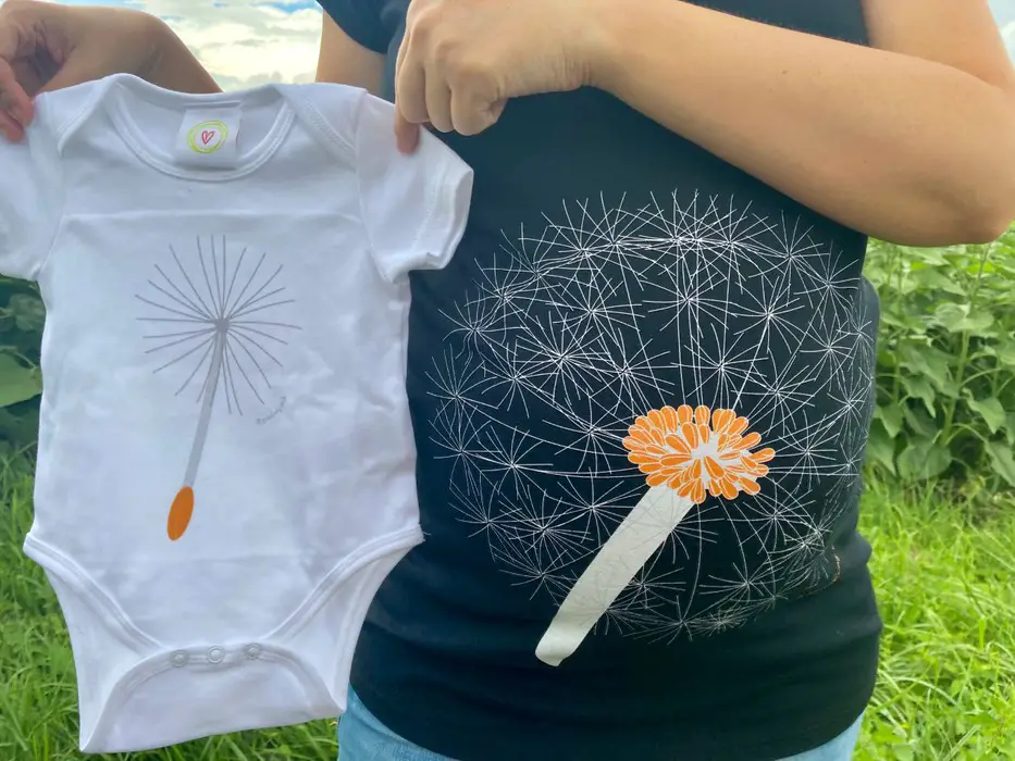 The photo shows a close up of my pregnancy bump with the dandelion design on the tshirt, I am also holding up a white baby grow that has a dandelion seed on it