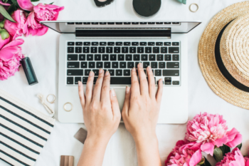 a laptop with a ladies hands typing. there are flowers, make up and jewellery around the laptop