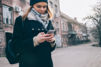 a lady stood by the side of a road looking at her phone, wearing a black woolen hat and coat with a grey scarf