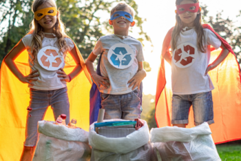 three children wearing superhero masks and capes and recycling signs on their tshirts as they stand next to recycling bags