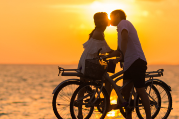 a man and lady kissing. they are by the sea, both holding onto their bikes with the sunset in the background