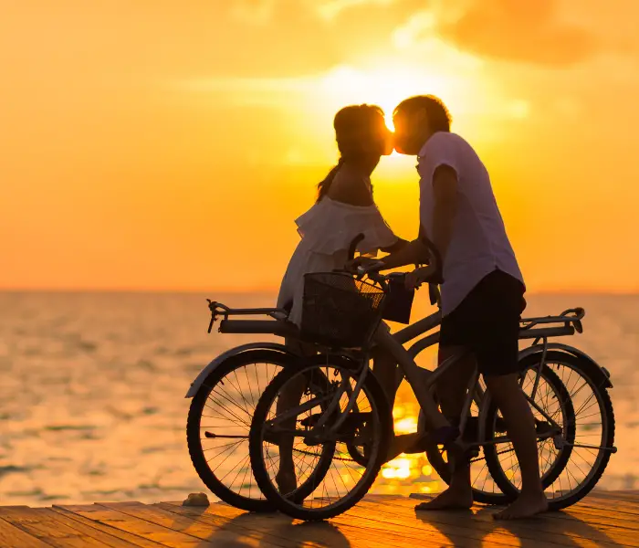 a man and lady kissing. they are by the sea, both holding onto their bikes with the sunset in the background