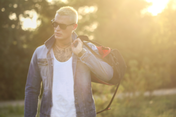 man wearing white tshirt, denim jacket and sunglasses, he is holding a bag over his shoulder.